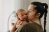 A photo of a woman with her hair in a plaited pony tail holding a new born. The woman is kissing the baby on the cheek and the baby is looking over her shoulder with their eyes open. 