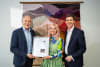 Dr Alan Finkel, Chairman of Proudly Human and Trevor Woods, CEO of Proudly Human present Maryanne Martin with her verification certificate in front of her tapestry, Wildflowers
