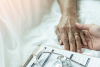 Close up image of hospital bed. Patient's being hold by another person. Doctor's clipboard and stethoscope in foreground. 